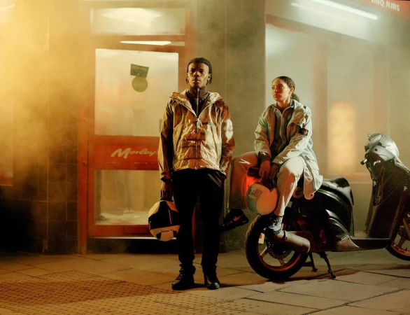 Two people pose in urban setting, motorcycle parked nearby. Man stands holding helmet, woman sits on motorcycle, wearing light-colored jackets.
