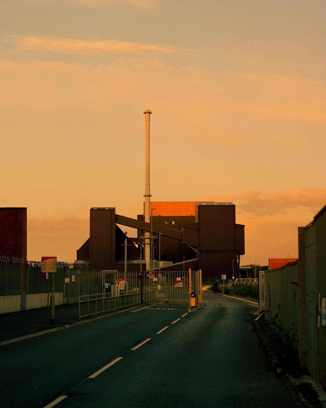 Industrial complex with smokestack and guardhouse, viewed from an empty road under a dusk sky with orange and purple hues.