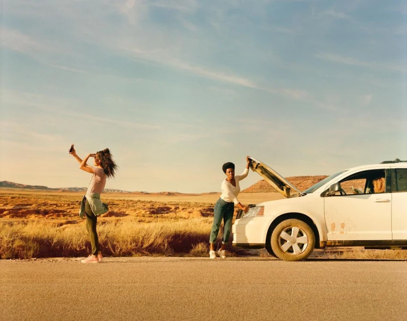 Two women on a desert road with a car. One woman uses a cell phone, the other repairs a car with the hood open on the side of a road.