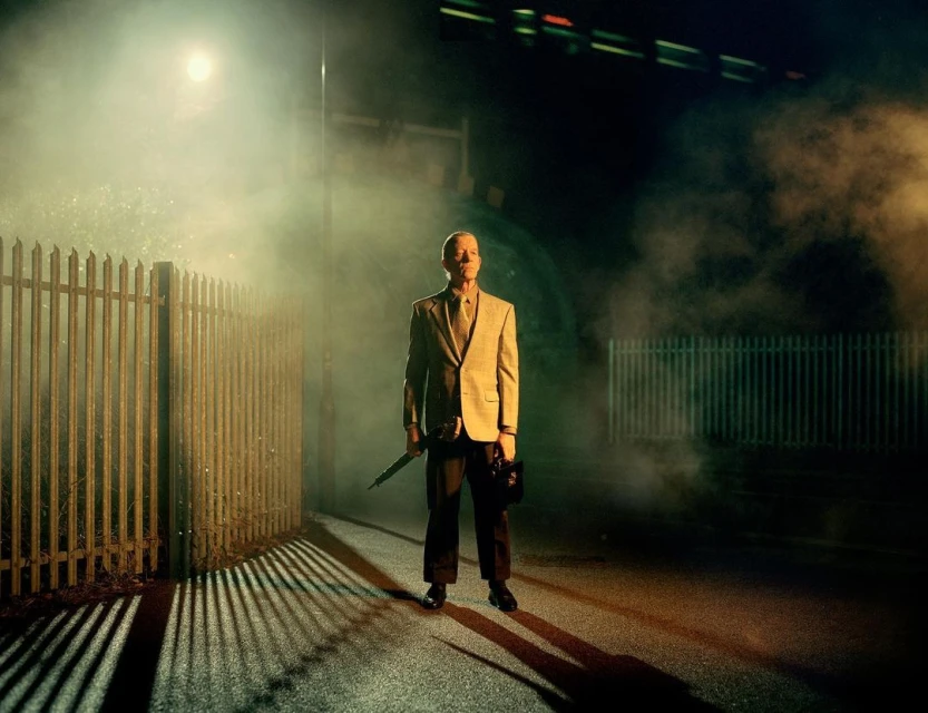 Mature man in suit stands on city street at night, illuminated by streetlight and diffused by fog, holding an umbrella and briefcase.