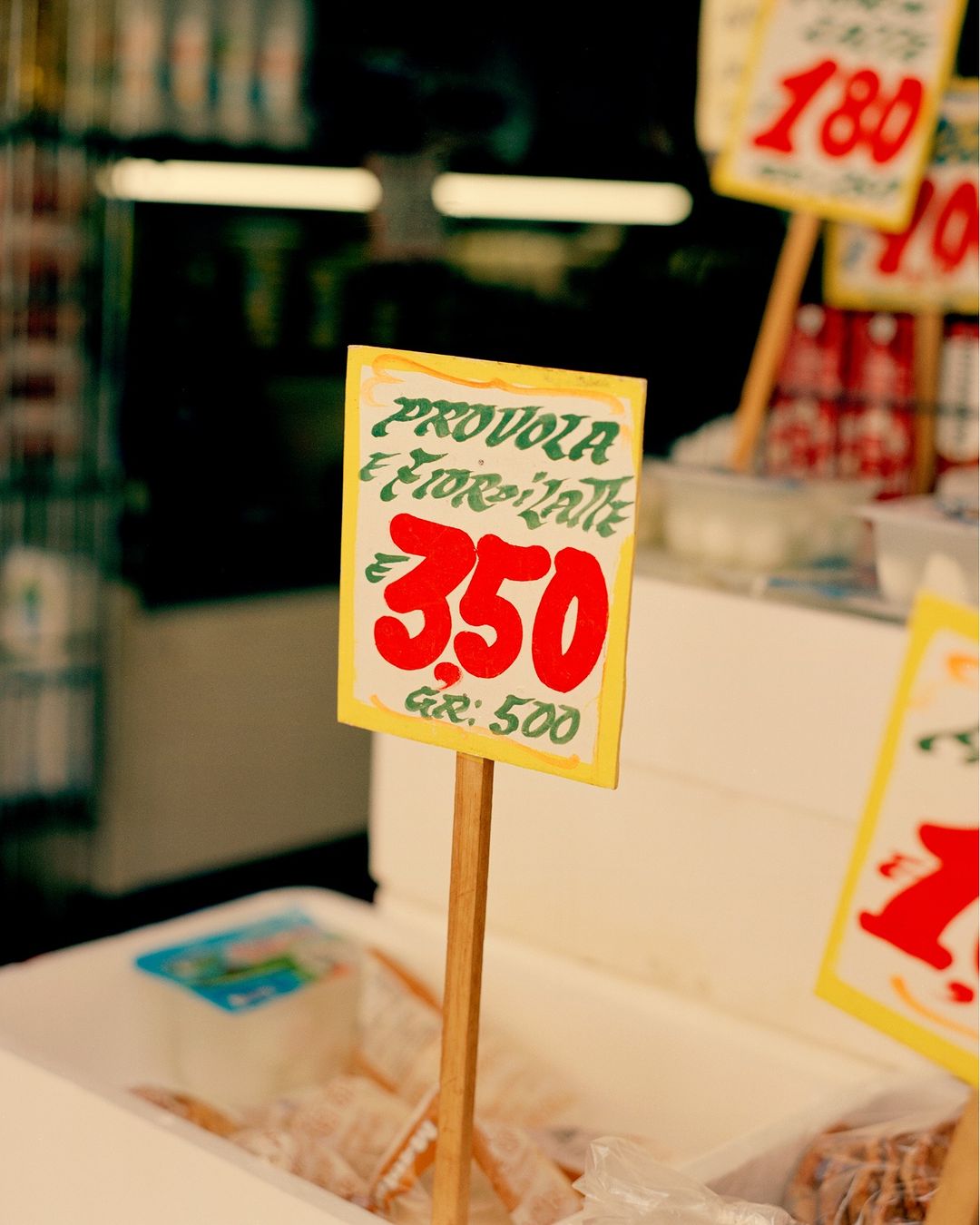 Close-up on handmade price signage for 'Provola e Fior di Latte' cheese in a deli, with red numbers on a yellow and green sign.