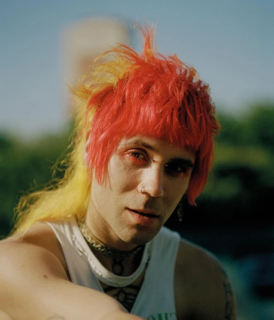 Close-up portrait of a young adult male with dyed red and yellow hair, a nose ring, and a tank top, posing outdoors during daylight.