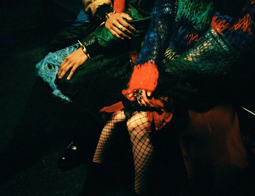 Close-up of a person wearing fishnet stockings, rings, and bracelets. They are wearing a colorful shag coat and black shoes in dark light.