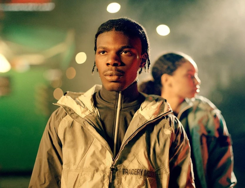 An adult Black male wearing a grey jacket stands in focus, with an adult Black female out of focus in the background, in an indoor setting.