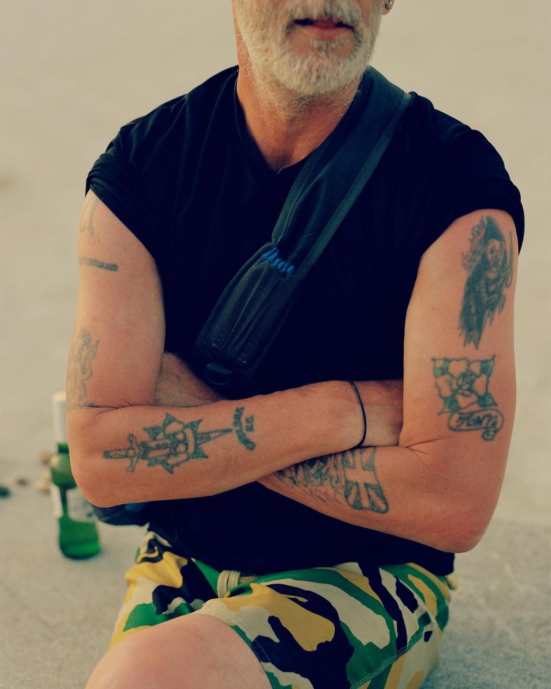 Adult male with arms crossed, wearing a black t-shirt and camouflage shorts, accessorized with a shoulder bag, showcasing arm tattoos.