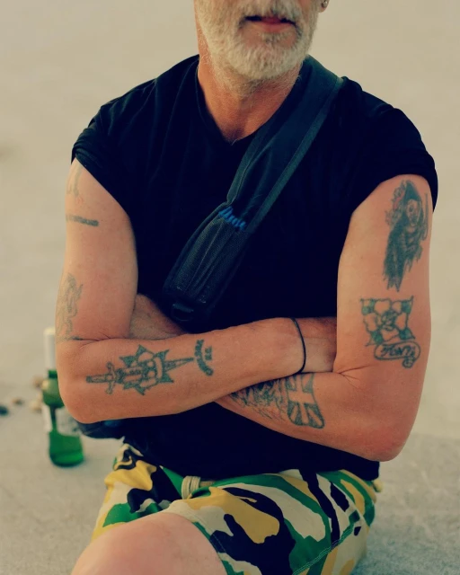 Adult male with arms crossed, wearing a black t-shirt and camouflage shorts, accessorized with a shoulder bag, showcasing arm tattoos.