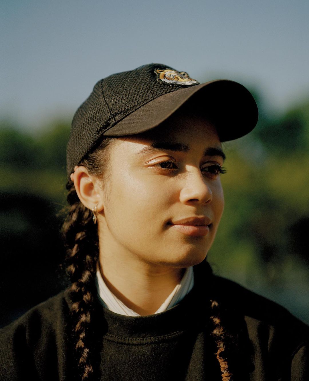 Close-up portrait of a young woman wearing a cap with braided hair looking to the side against a blurred outdoor background.