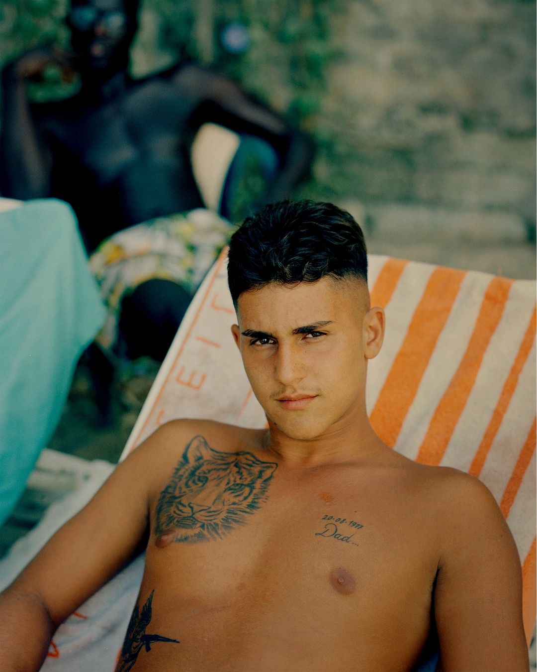 Young man lounges on a striped beach chair with chest tattoos, while another man relaxes in the background under the sunlight.