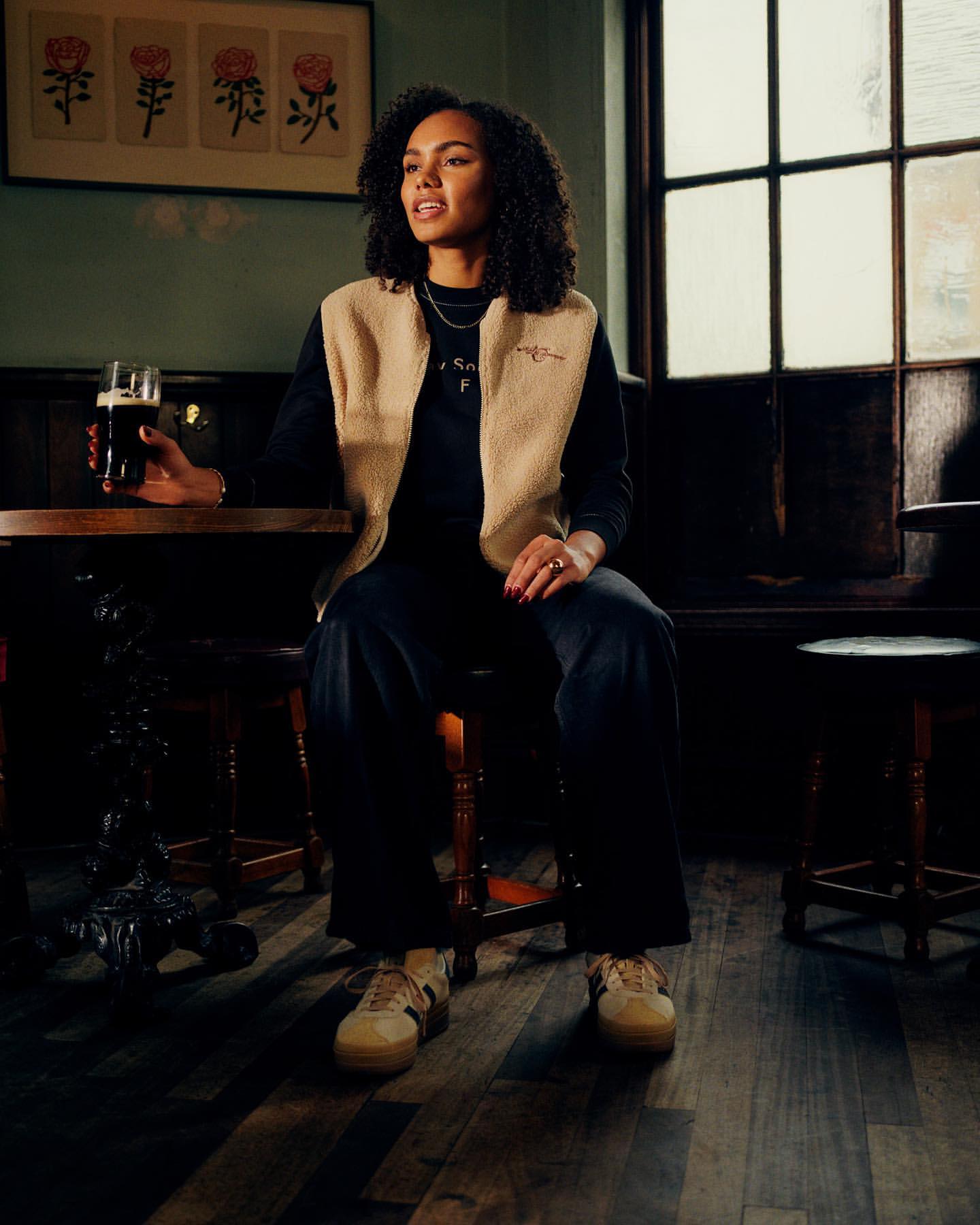 Woman in vest seated at a wooden table with a drink in a bar, with floral artwork on the wall and natural light from the window.