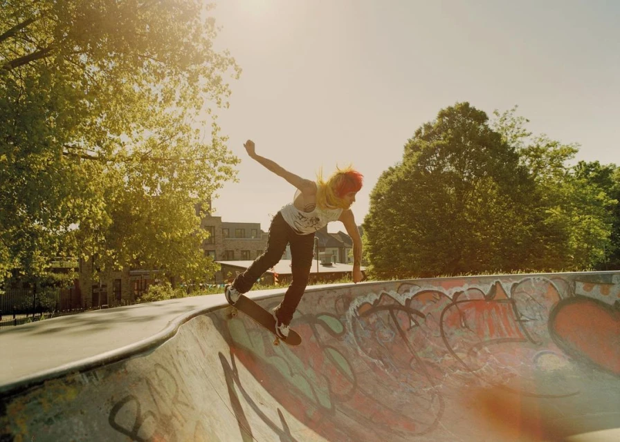 A skateboarder on a ramp in an urban skatepark under bright sunlight, surrounded by trees and a building, capturing an active outdoor scene.