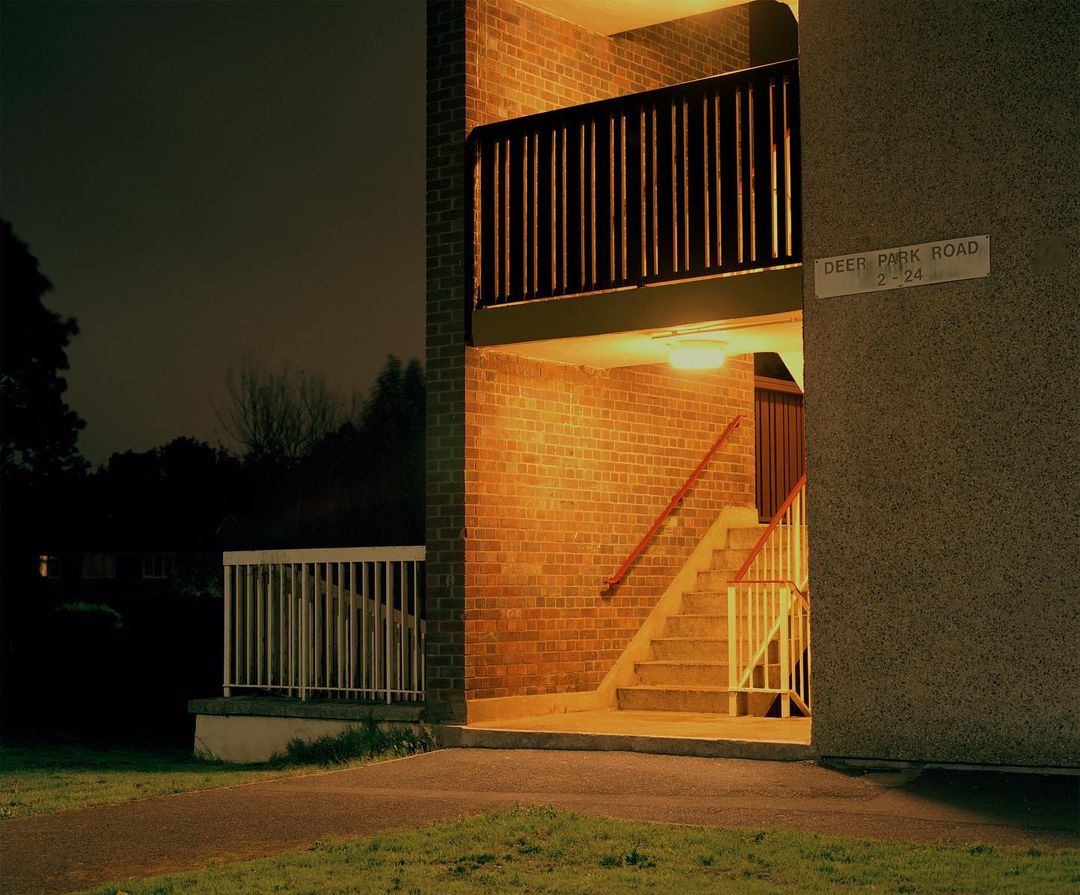 Exterior of an illuminated brick apartment building at night with stairs leading up, featuring warm lighting and architectural details.