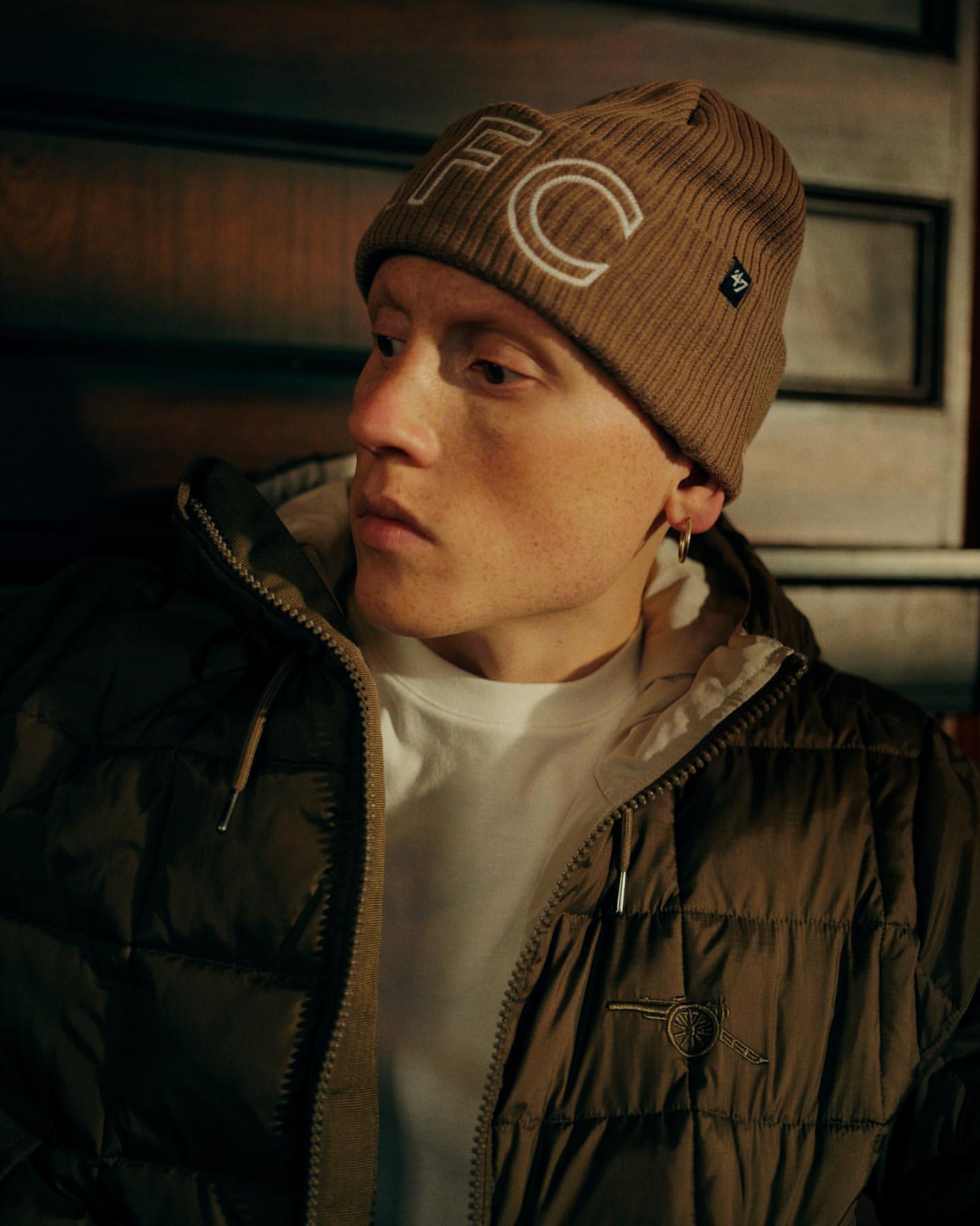 Portrait of a young man wearing a brown beanie with white FC letters, a white shirt, and a puffy olive-green jacket with a zipper.