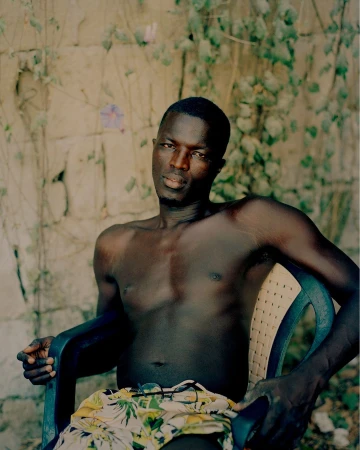 Shirtless Black man seated outdoors on a plastic patio chair in front of a rustic stone wall and green foliage.