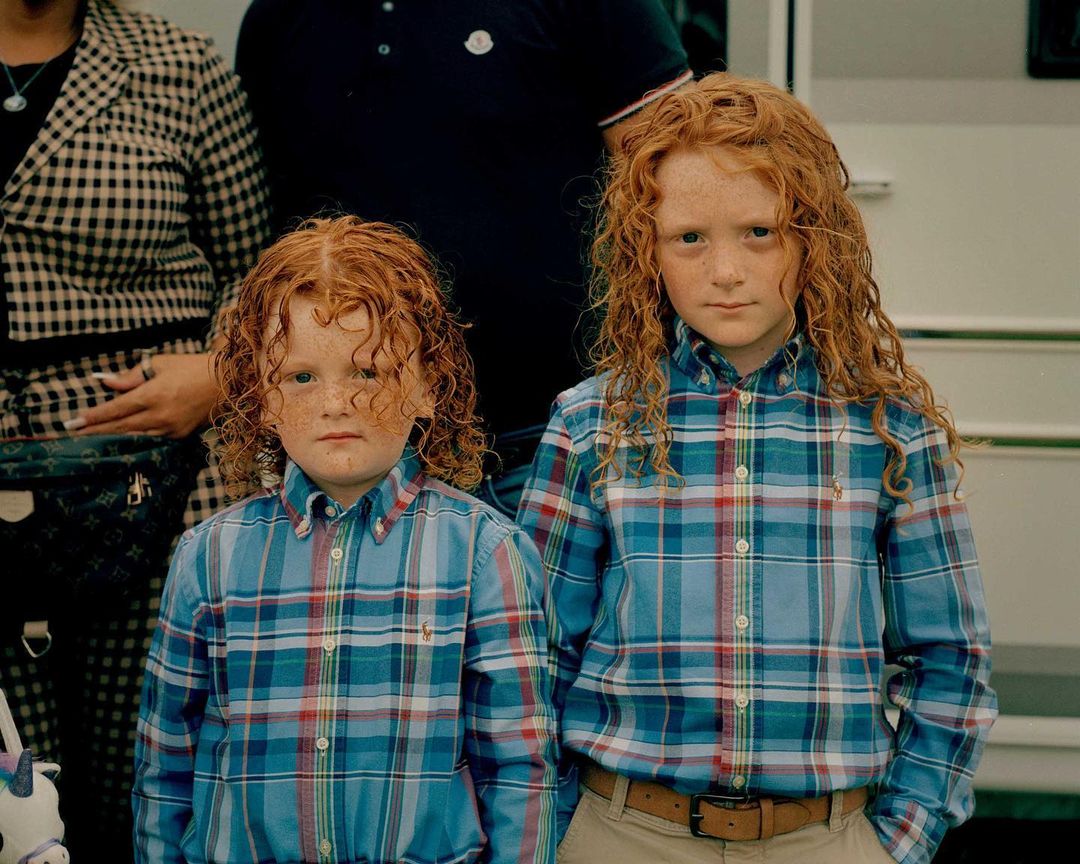 Two red-haired children in plaid shirts pose in front of a white trailer, with adults visible in the background. Outdoor setting.