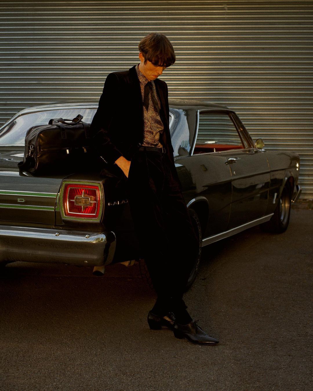 An adult male wearing a suit leans against a black vintage car parked outside a building with metal siding, illuminated by sunlight.