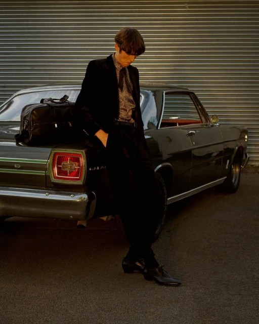 An adult male wearing a suit leans against a black vintage car parked outside a building with metal siding, illuminated by sunlight.