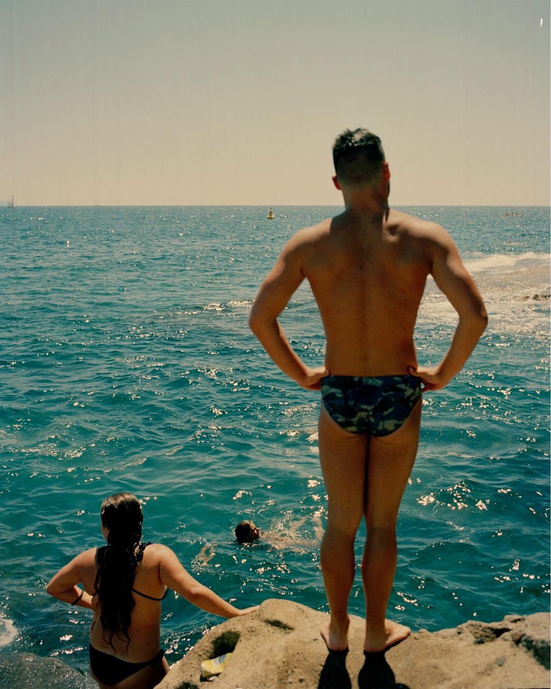 Three people enjoy a sunny day at the beach, with a man standing on a rock overlooking the ocean and others swimming in the water.