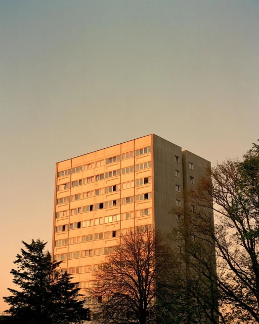 High-rise apartment building rises prominently in an urban setting, framed by silhouetted trees against a light blue and orange sky at dusk.