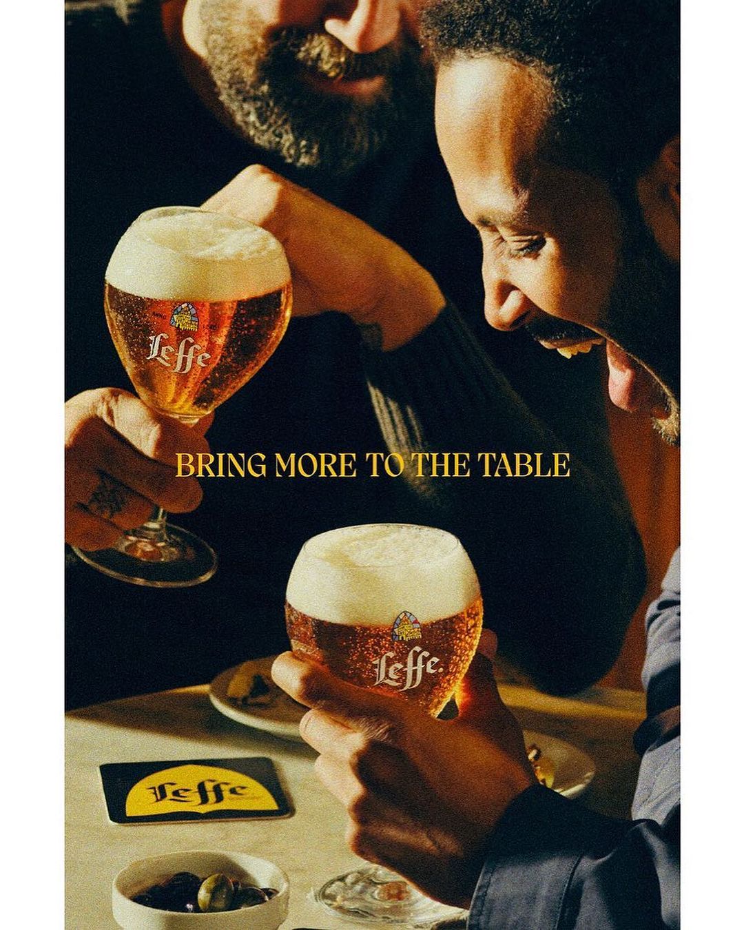 Two adult males at a table holding Leffe beer glasses filled with beer, offering a toast; Leffe logo on glass and coaster is visible.