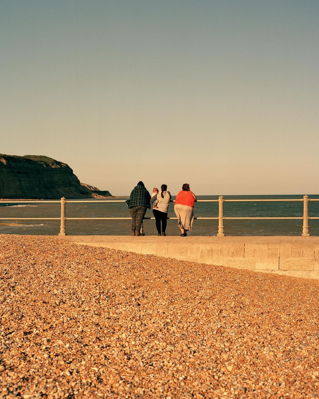Three people stand on a concrete walkway along a coast with a metal railing, overlooking the ocean on a sunny day with a cliff.