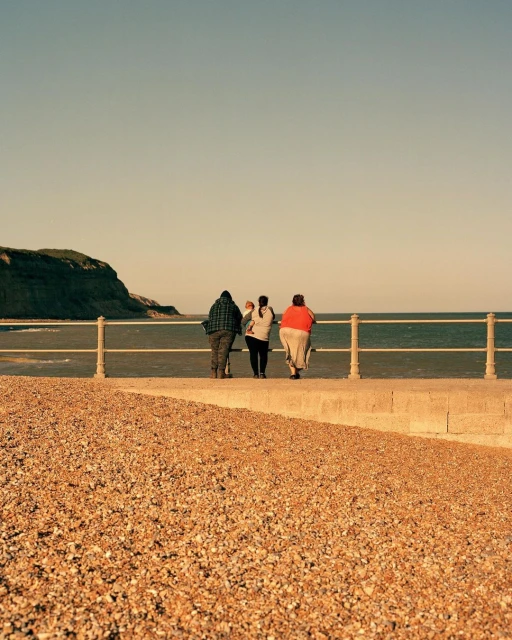 Three people stand on a concrete walkway along a coast with a metal railing, overlooking the ocean on a sunny day with a cliff.