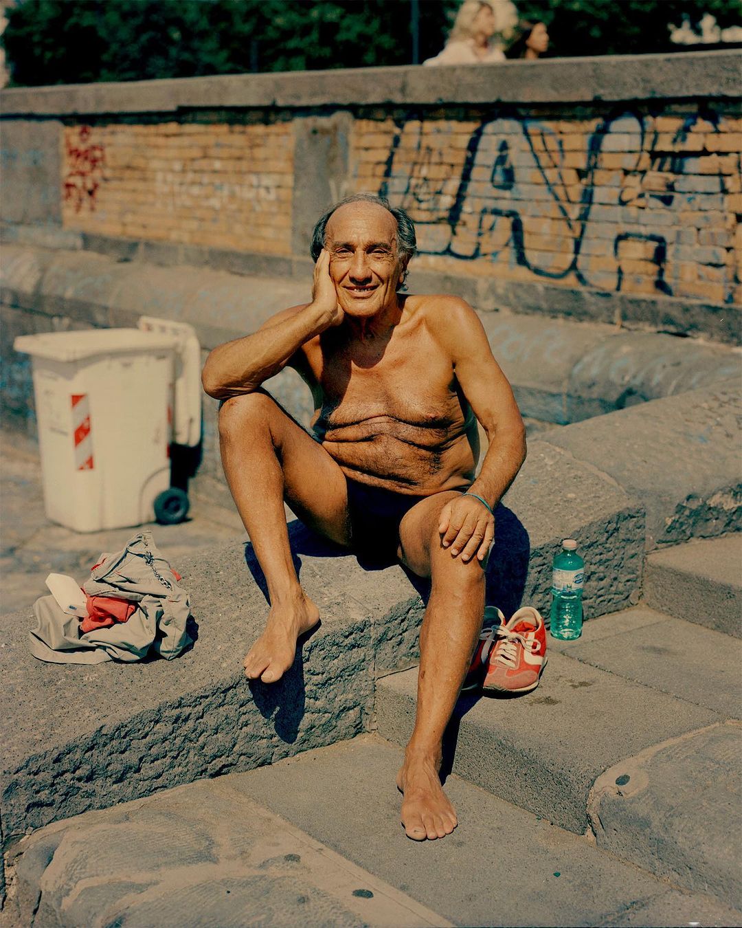 Elderly man seated outdoors on concrete steps wearing dark swim briefs with a water bottle and sneakers nearby.