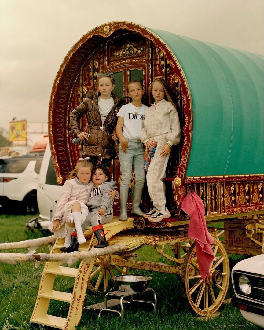 A group of five children poses in front of an ornate, green and gold wagon in a grassy field with cars visible in the distance.