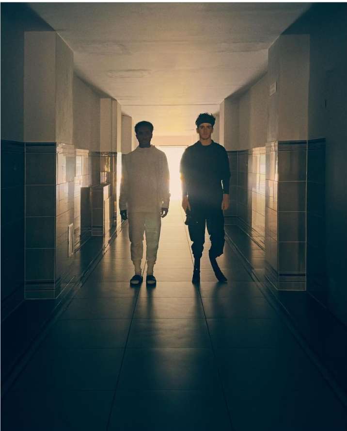 Two men standing inside a long tiled hallway against a bright light source at the far end by Sam Wright
