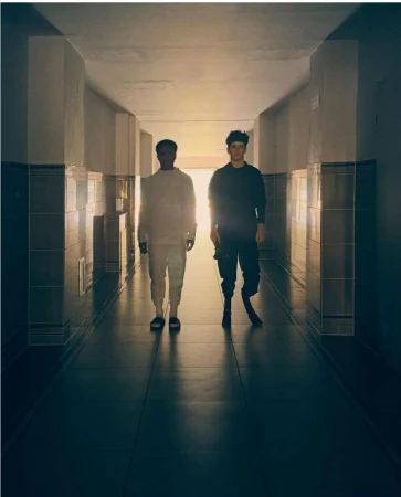 Two men standing inside a long tiled hallway against a bright light source at the far end by Sam Wright