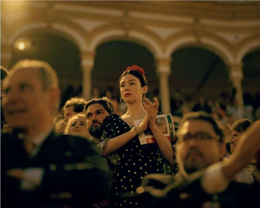 Indoor shot of a diverse audience watching a show; a woman in a polka dot dress and flower hairpiece claps appreciatively.