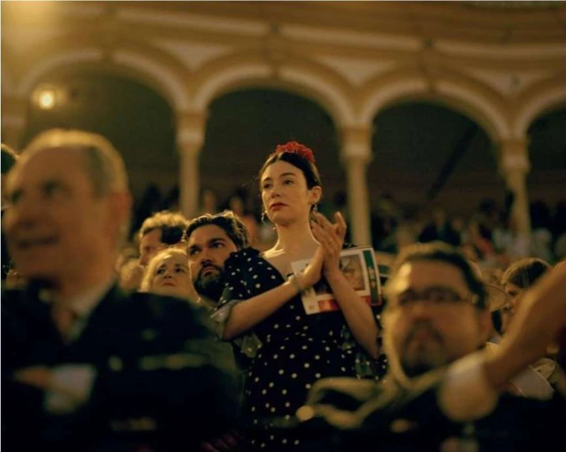 Indoor shot of a diverse audience watching a show; a woman in a polka dot dress and flower hairpiece claps appreciatively.