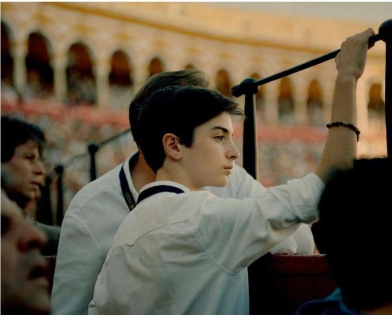 A teen boy in a white shirt stands inside an arena with multiple people and architectural arches, facing right and holding onto a metal rail.