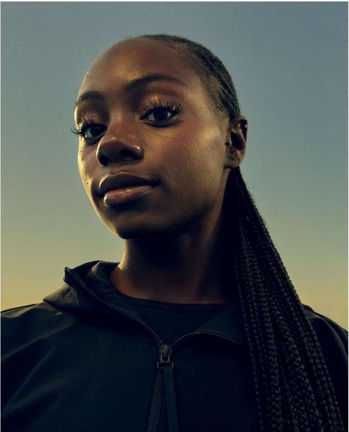 Close-up portrait of a young Black woman with braided hair, wearing a dark zip-up jacket, set against a gradient sky background.