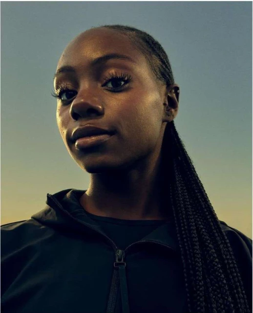 Close-up portrait of a young Black woman with braided hair, wearing a dark zip-up jacket, set against a gradient sky background.