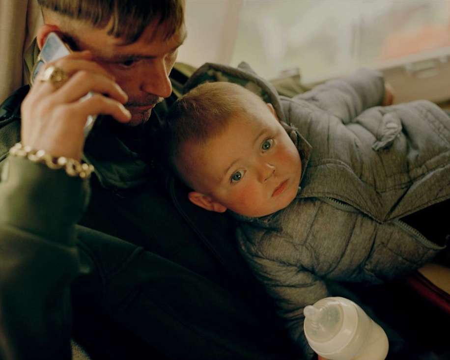 Man holding baby in grey quilted jacket while talking on smartphone. Father and child inside with baby bottle in hand.