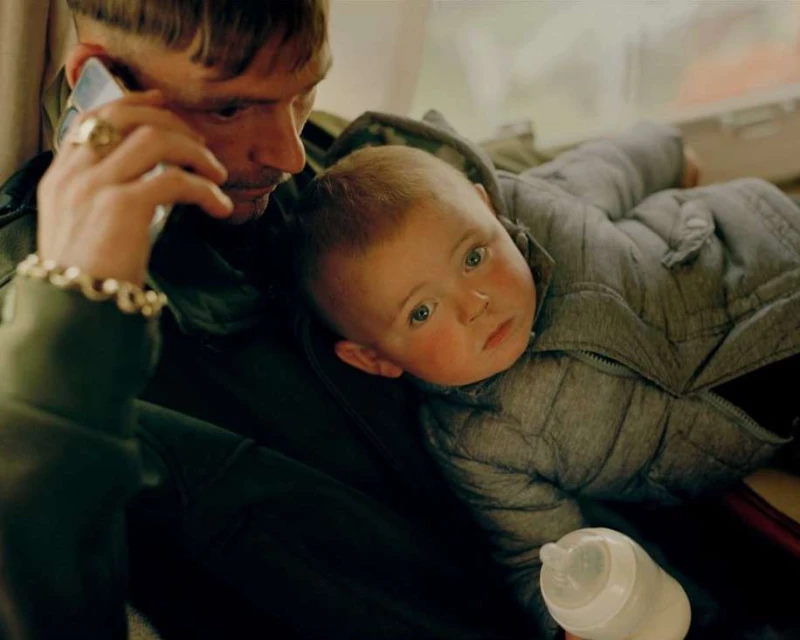 Man holding baby in grey quilted jacket while talking on smartphone. Father and child inside with baby bottle in hand.