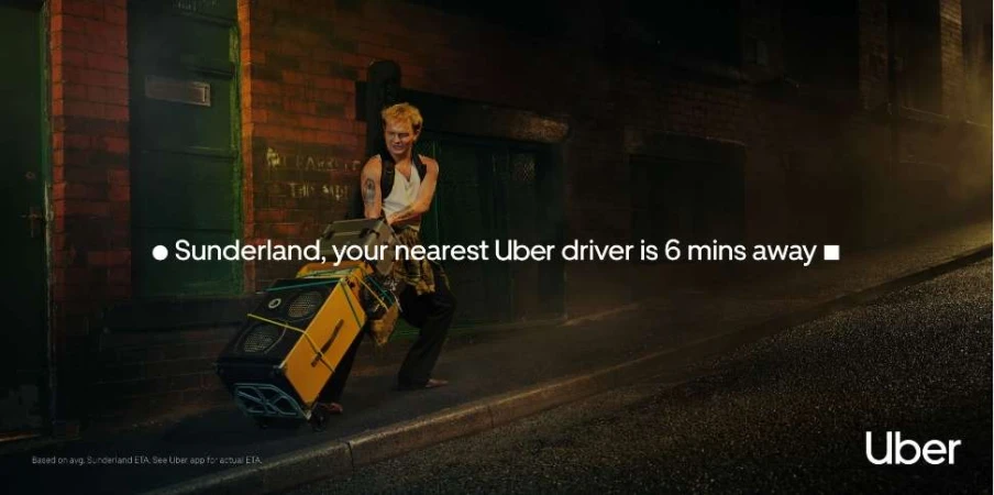 Night scene shows a man with a sound system on wheels walking on a city street; Uber ad, brick building & dark urban background.