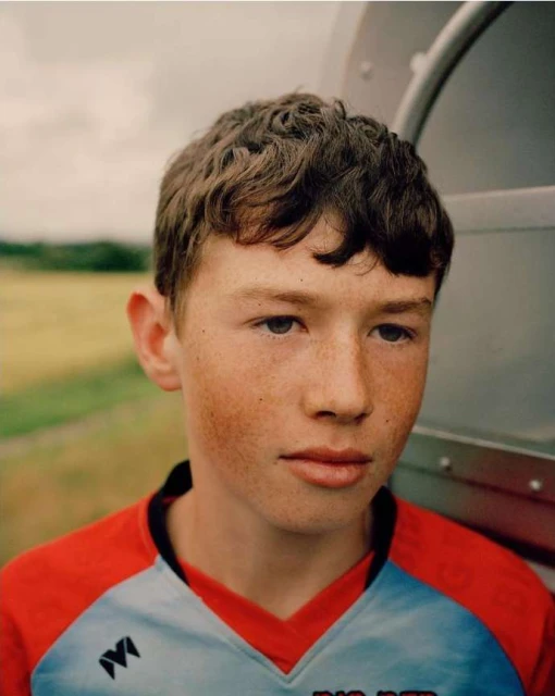 Portrait of a young person with freckles, wearing a red and light-blue shirt, standing outdoors with a cloudy sky in the background.