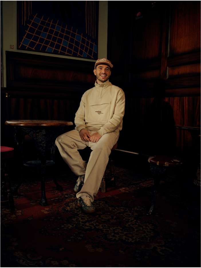 Man sitting on a stool in an indoor space, wearing a beige tracksuit and hat, with a patterned rug on the floor.