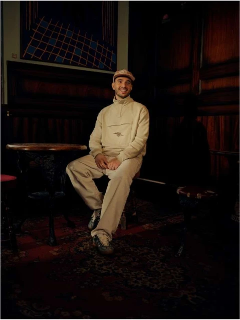 Man sitting on a stool in an indoor space, wearing a beige tracksuit and hat, with a patterned rug on the floor.