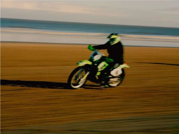 Action shot of a motocross rider on a green and white dirt bike speeding across a sandy beach with the ocean in the background.