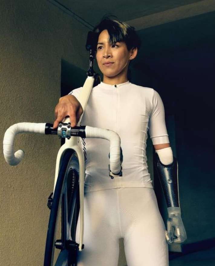 Adult female with prosthetic arm poses with a white track bicycle, indoors with dark walls in a medium, eye-level shot.