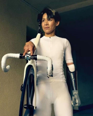 Adult female with prosthetic arm poses with a white track bicycle, indoors with dark walls in a medium, eye-level shot.