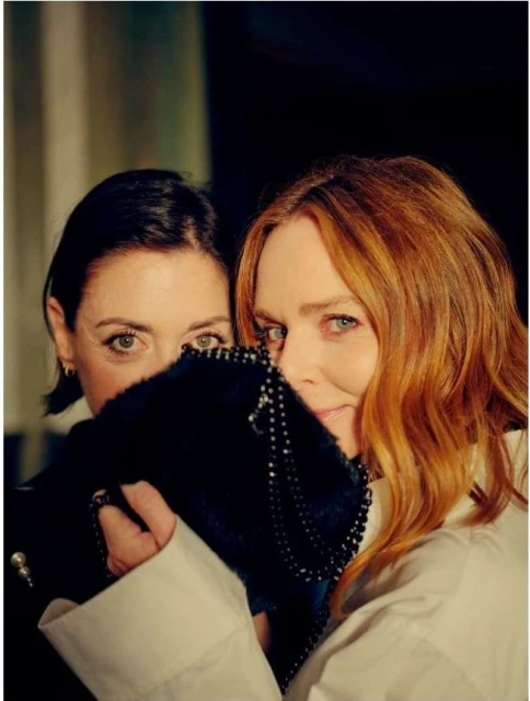 Two adult women pose indoors, one partially hidden by a beaded black bag, against a dark backdrop with subtle lighting and neutral tones.