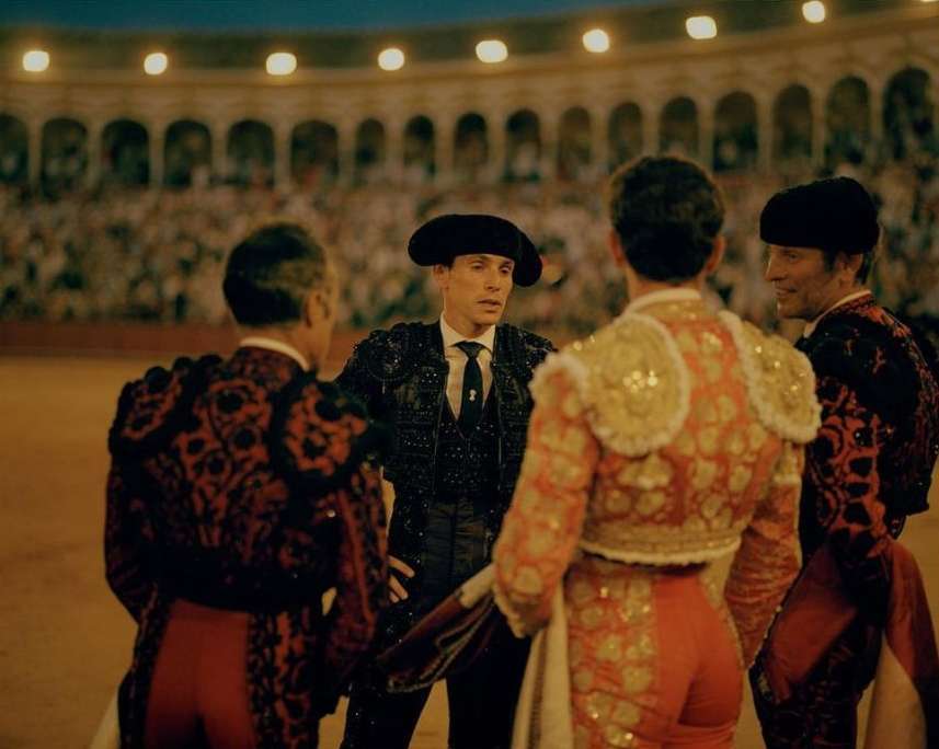 Four bullfighters in ornate costumes gather in an arena, captured in a medium shot with a crowd visible in the background.