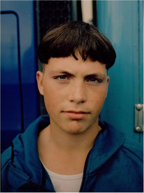 A young person with a bowl cut hairstyle gazes directly at the camera, wearing a blue hooded sweatshirt against a blue background.