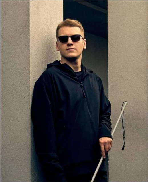 Man with blonde hair and sunglasses wearing a black jacket stands beside a grey wall with a white cane in an outdoor setting.