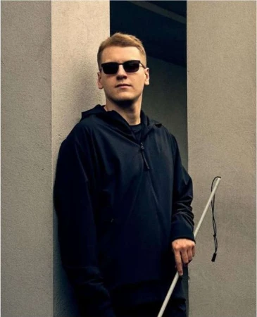 Man with blonde hair and sunglasses wearing a black jacket stands beside a grey wall with a white cane in an outdoor setting.