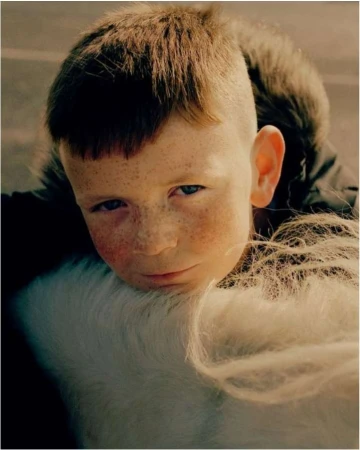 Close-up portrait of a young boy with freckles resting his chin on a white horse mane, photographed by Sam Wright.