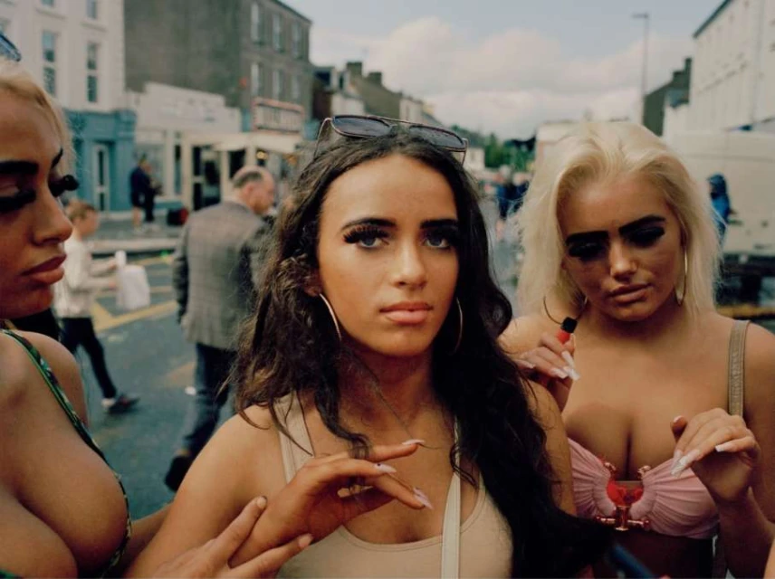 Three young women pose in an urban setting. The woman in the center wears sunglasses on her head and a light-colored tank top.